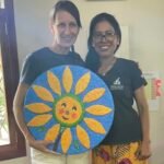 two women holding a round drawing showing a yellow flower Letty