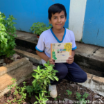 niño agachado junto a la planta Letty sosteniendo un libro de trabajo